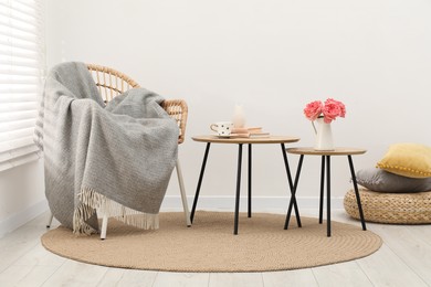 Photo of Wicker chair with gray woolen blanket, books, beautiful flowers in vase and cup on nesting tables indoors
