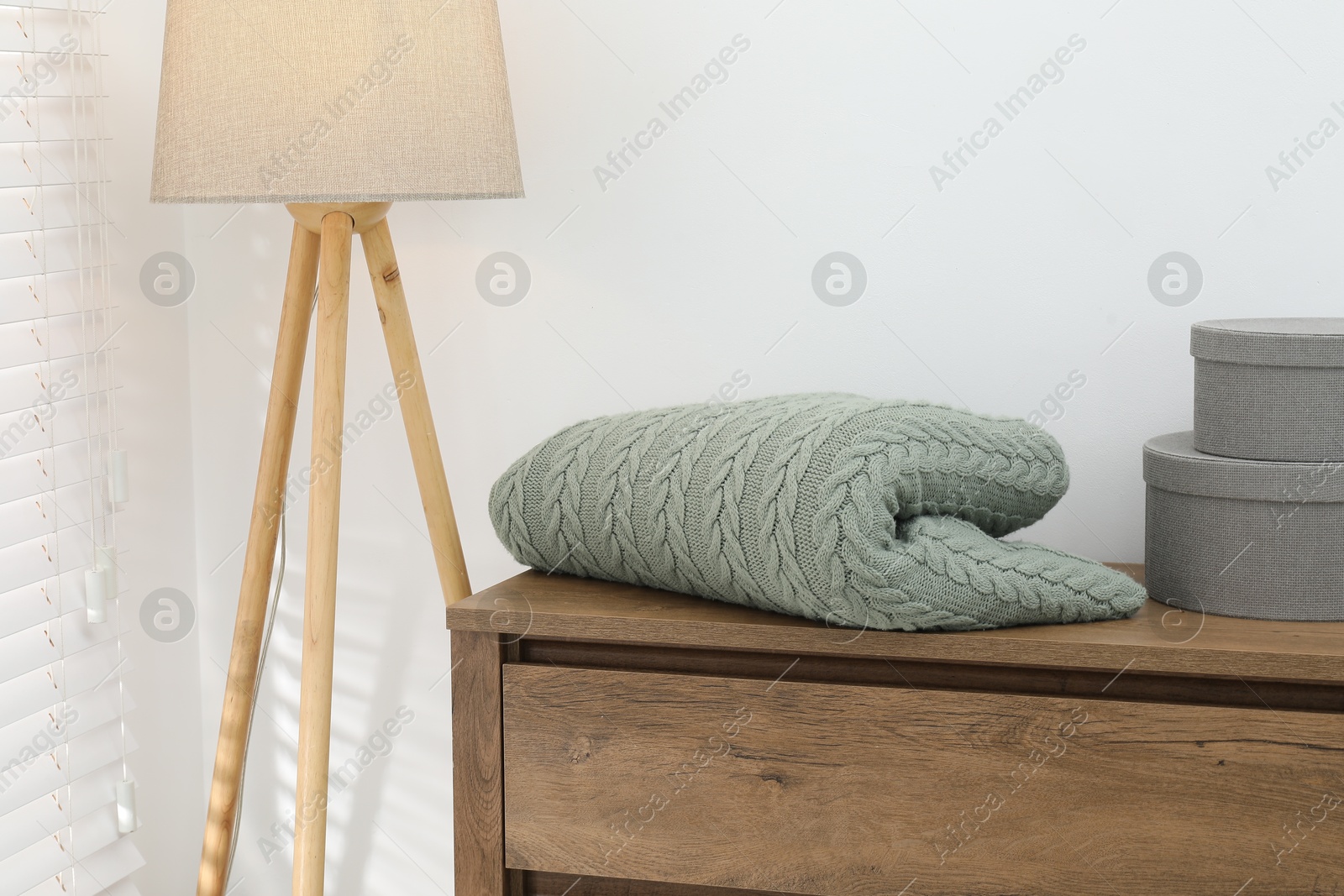 Green woolen blanket, boxes on wooden chest of drawers and lamp near white wall indoors, closeup Photo of Green woolen blanket, boxes on wooden chest of drawers and lamp near white wall indoors, closeup
