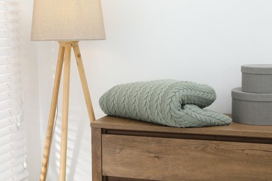 Photo of Green woolen blanket, boxes on wooden chest of drawers and lamp near white wall indoors, closeup