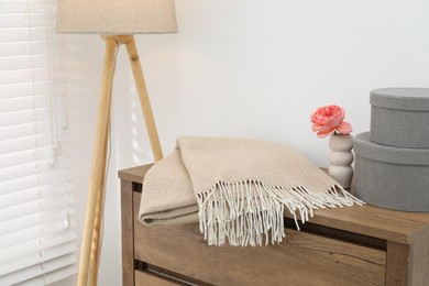 Photo of Beige woolen blanket, boxes, beautiful flower in vase on wooden chest of drawers and lamp near white wall indoors, closeup