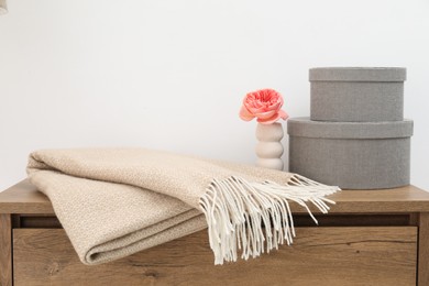 Photo of Woolen blanket, boxes and beautiful flower in vase on wooden chest near white wall indoors, closeup