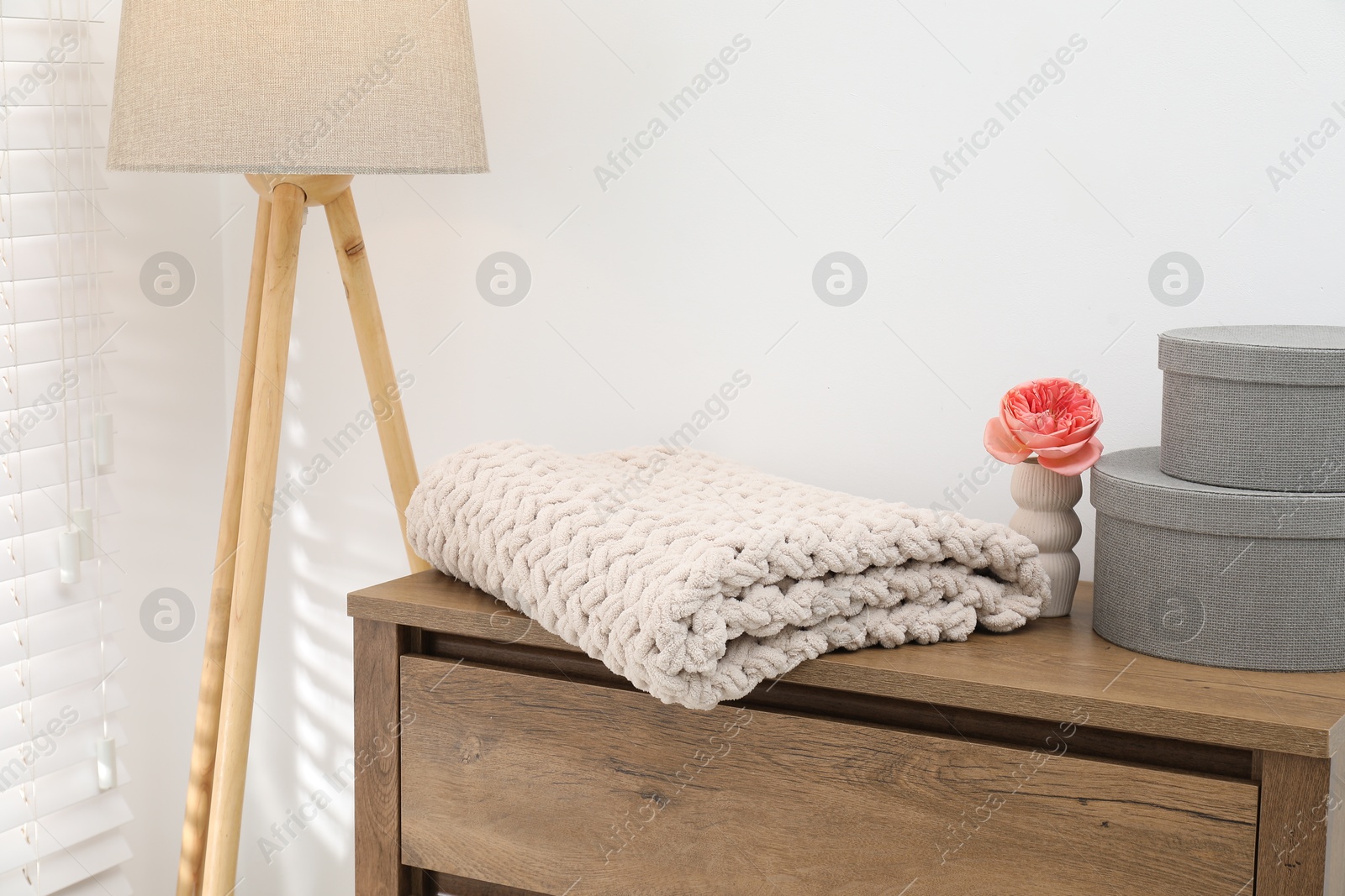 Woolen blanket, boxes, beautiful flower in vase on wooden chest of drawers and lamp near white wall indoors, closeup Photo of Woolen blanket, boxes, beautiful flower in vase on wooden chest of drawers and lamp near white wall indoors, closeup