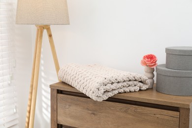Photo of Woolen blanket, boxes, beautiful flower in vase on wooden chest of drawers and lamp near white wall indoors, closeup