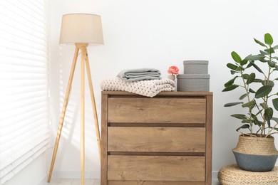 Photo of Woolen blankets on wooden chest of drawers, lamp and houseplant near white wall indoors
