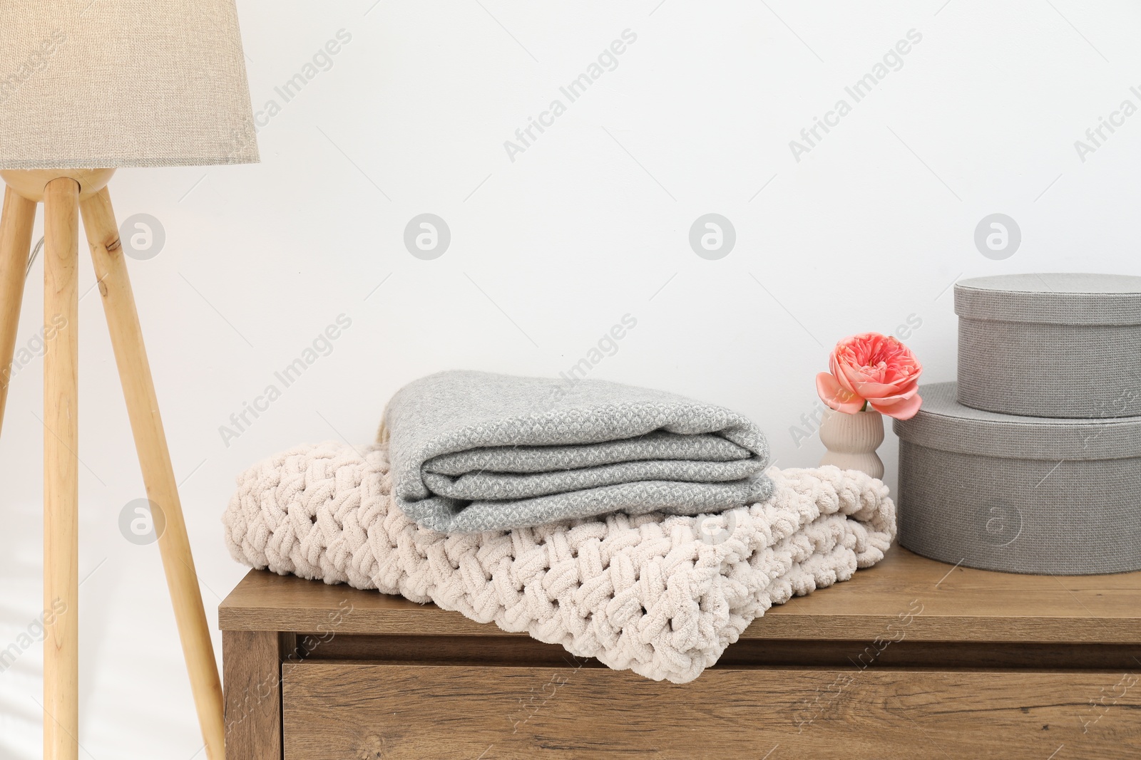 Woolen blankets, beautiful flower in vase and boxes on wooden chest of drawers near white wall indoors, closeup Photo of Woolen blankets, beautiful flower in vase and boxes on wooden chest of drawers near white wall indoors, closeup