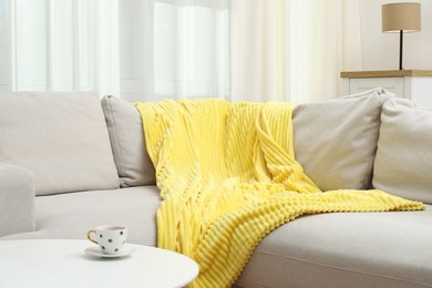 Photo of Comfortable sofa with yellow woolen blanket and cup on table indoors