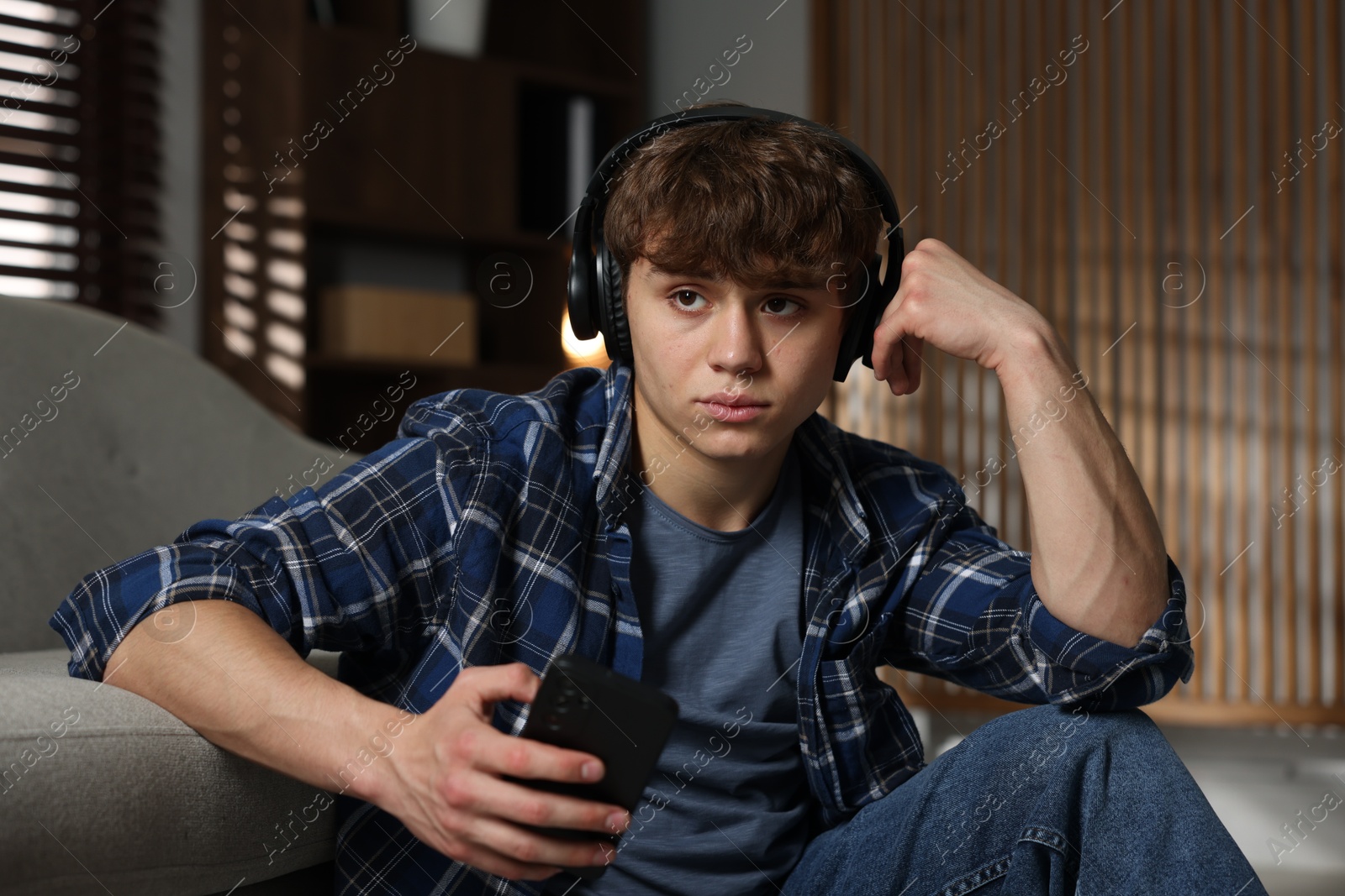Teen problems. Upset teenage boy in headphones with smartphone near sofa at home Photo of Teen problems. Upset teenage boy in headphones with smartphone near sofa at home