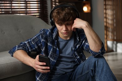 Photo of Teen problems. Upset teenage boy in headphones with smartphone near sofa at home