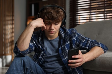 Photo of Teen problems. Upset teenage boy in headphones with smartphone near sofa at home