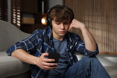 Photo of Teen problems. Upset teenage boy in headphones with smartphone near sofa at home