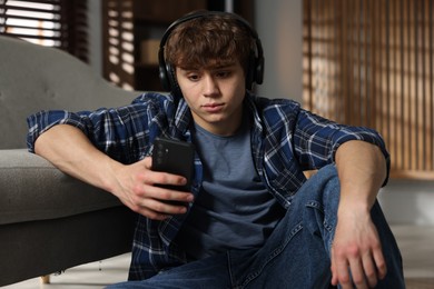 Photo of Teen problems. Upset teenage boy in headphones with smartphone near sofa at home
