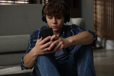 Photo of Teen problems. Upset teenage boy in headphones with smartphone near sofa at home