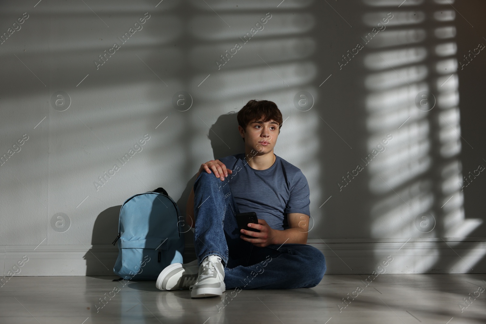 Teen problems. Upset teenage boy with smartphone and backpack near light wall indoors Photo of Teen problems. Upset teenage boy with smartphone and backpack near light wall indoors