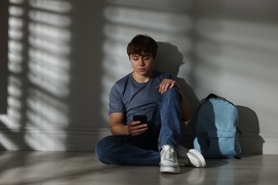 Photo of Teen problems. Upset teenage boy with smartphone and backpack near light wall indoors