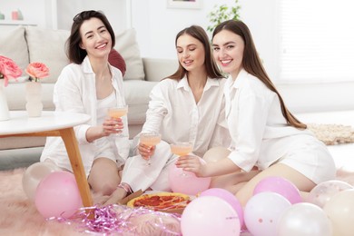 Photo of Happy friends in pajamas with glasses of alcoholic drink eating pizza on floor at home