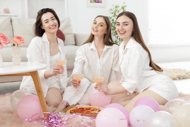 Photo of Happy friends in pajamas with glasses of alcoholic drink eating pizza on floor at home