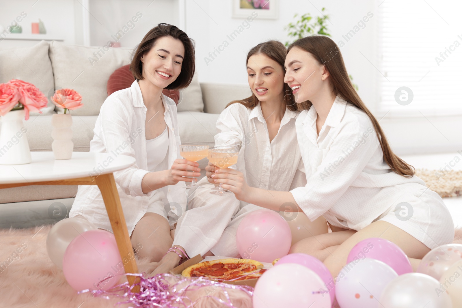 Happy friends in pajamas clinking glasses of alcoholic drink near pizza on floor at home Photo of Happy friends in pajamas clinking glasses of alcoholic drink near pizza on floor at home