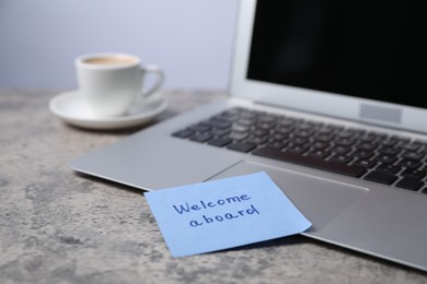 Photo of Sticky note with phrase Welcome Aboard, laptop and coffee on grey table, closeup