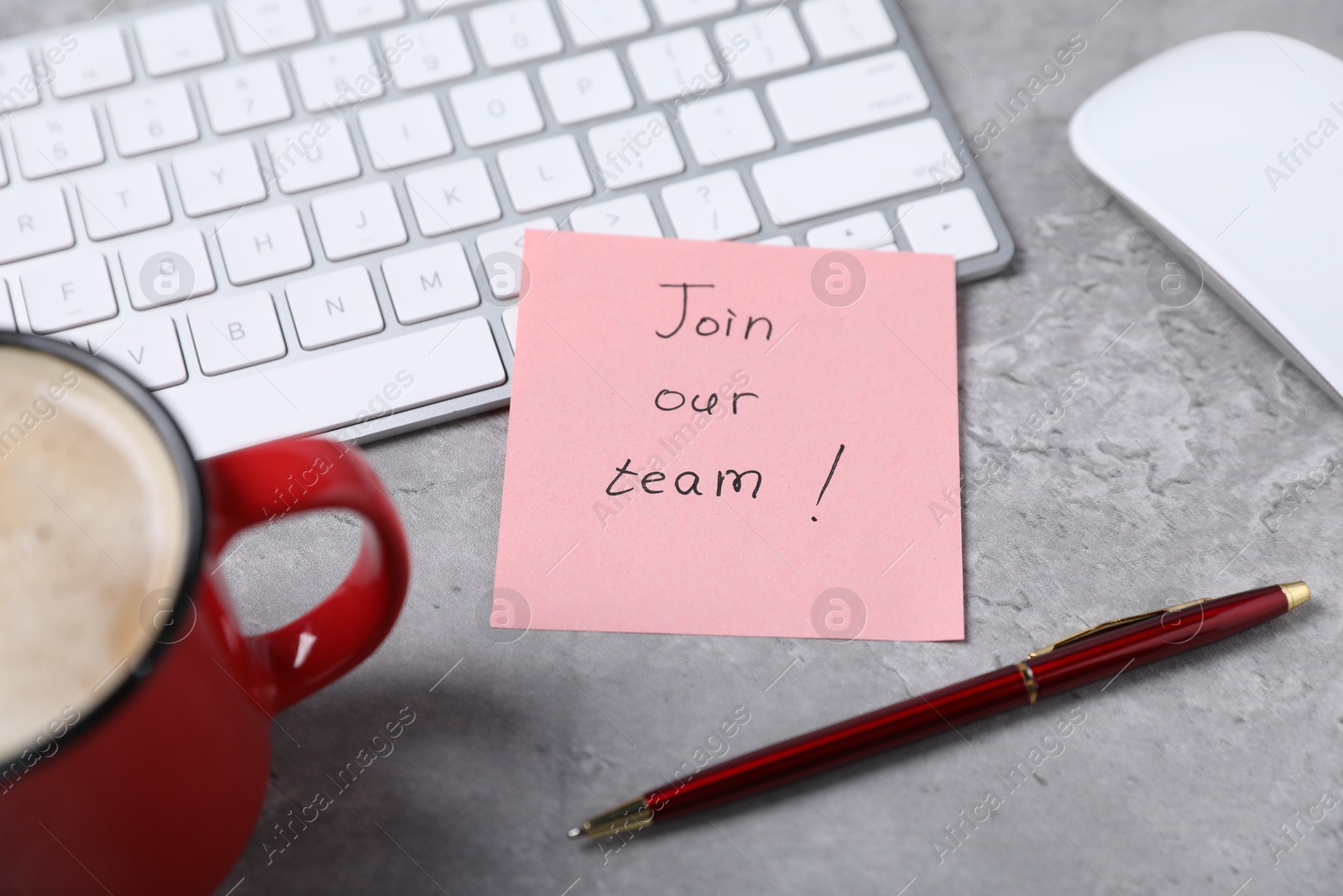 Photo of Sticky note with phrase Join Our Team, computer keyboard, mouse, pen and coffee on grey table, closeup