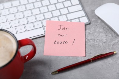 Photo of Sticky note with phrase Join Our Team, computer keyboard, mouse, pen and coffee on grey table, closeup
