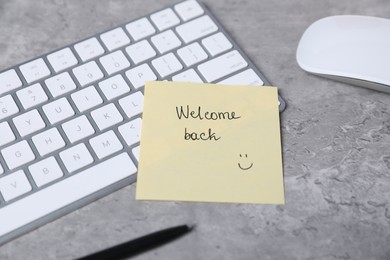 Photo of Sticky note with phrase Welcome Back, computer keyboard, mouse and pen on grey table, closeup