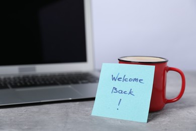 Photo of Sticky note with phrase Welcome Back, coffee and laptop on grey table, closeup