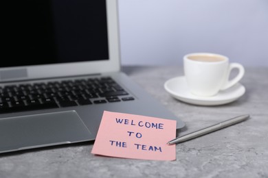 Photo of Sticky note with phrase Welcome To The Team, coffee, pen and laptop on grey table, closeup