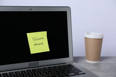 Photo of Sticky note with phrase Welcome Aboard, coffee and laptop on grey table, closeup