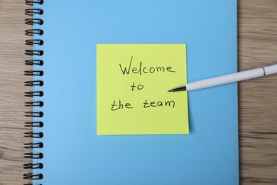 Photo of Sticky note with phrase Welcome To The Team, pen and notebook on wooden table, top view