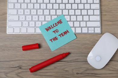 Photo of Sticky note with phrase Welcome To The Team, marker, computer keyboard and mouse on wooden table, flat lay
