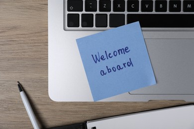 Photo of Sticky note with phrase Welcome Aboard, laptop and pen on wooden table, flat lay