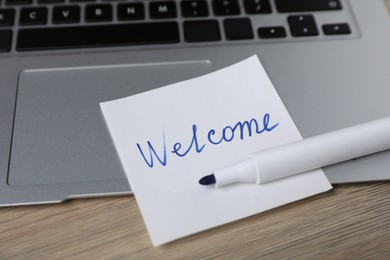Photo of Sticky note with word Welcome, laptop and marker on wooden table, closeup
