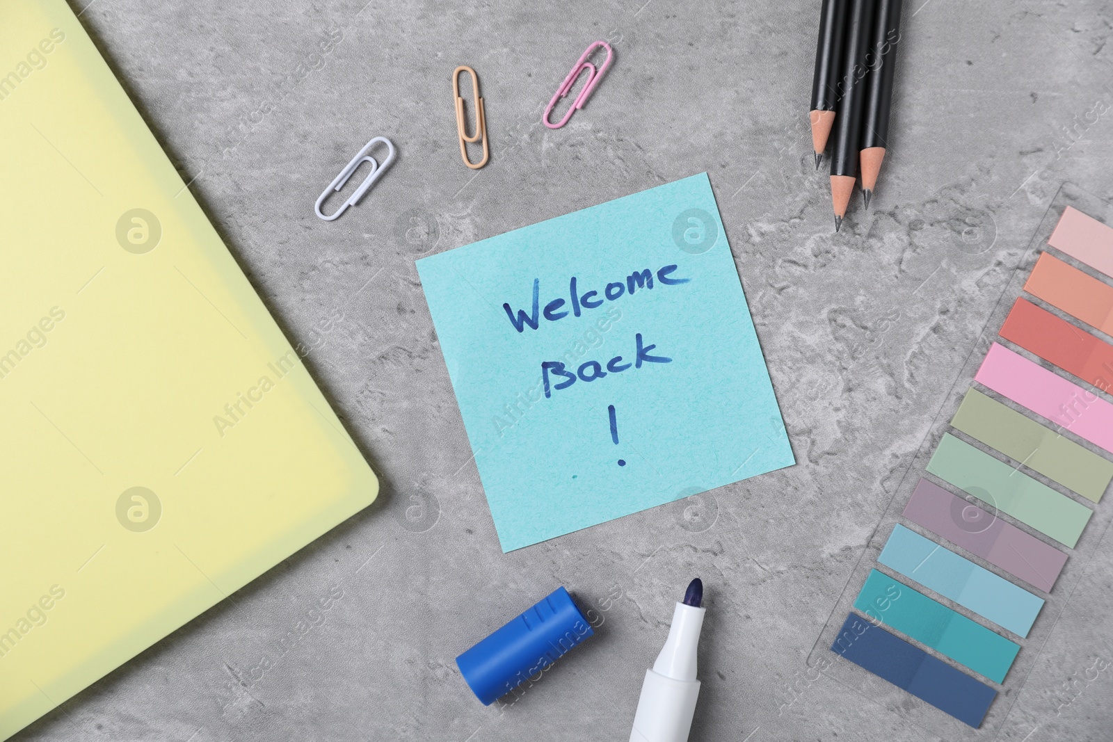 Sticky note with phrase Welcome Back and different stationery on grey table, flat lay Photo of Sticky note with phrase Welcome Back and different stationery on grey table, flat lay