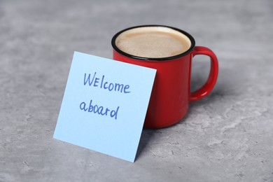 Photo of Sticky note with phrase Welcome Aboard and coffee on grey table, closeup