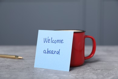Photo of Sticky note with phrase Welcome Aboard, coffee and pen on grey table, closeup
