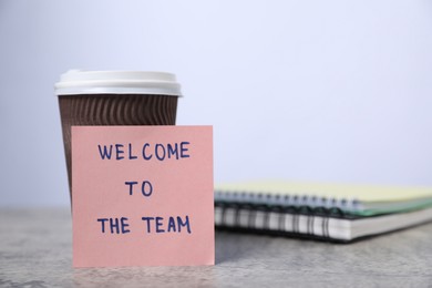 Photo of Sticky note with phrase Welcome To The Team, coffee and notebooks on grey table, closeup
