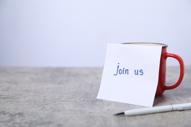 Photo of Sticky note with phrase Join Us, coffee and pen on grey table, closeup. Space for text