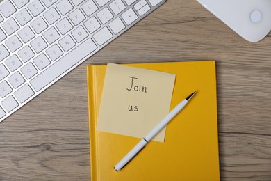 Photo of Sticky note with phrase Join Us, pen, notebook, computer keyboard and mouse on wooden table, flat lay