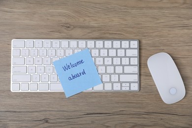 Photo of Sticky note with phrase Welcome Aboard, computer keyboard and mouse on wooden table, flat lay