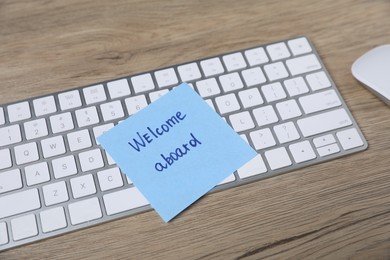 Photo of Sticky note with phrase Welcome Aboard, computer keyboard and mouse on wooden table, closeup