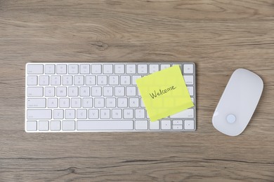 Photo of Sticky note with word Welcome, computer keyboard and mouse on wooden table, flat lay