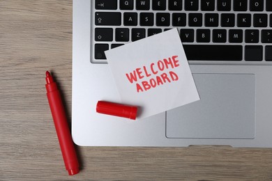 Photo of Sticky note with phrase Welcome Aboard, laptop and marker on wooden table, flat lay