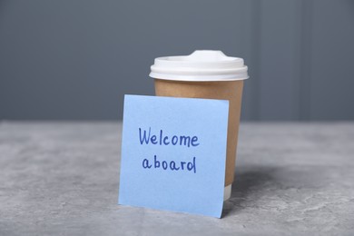 Photo of Sticky note with phrase Welcome Aboard and coffee on grey table, closeup