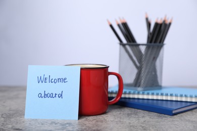 Photo of Sticky note with phrase Welcome Aboard, coffee, notebooks and pencils on grey table, closeup