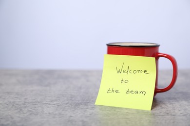 Photo of Sticky note with phrase Welcome To The Team and coffee on grey table, closeup. Space for text