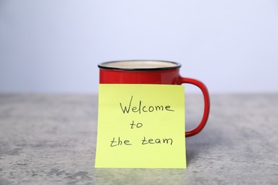 Photo of Sticky note with phrase Welcome To The Team and coffee on grey table, closeup