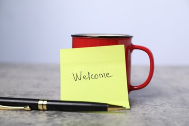 Photo of Sticky note with word Welcome, coffee and pen on grey table, closeup