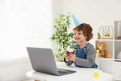 Photo of Birthday celebration. Little boy with cupcake having video call via laptop at table indoors. Space for text