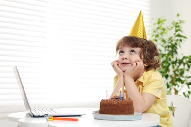 Photo of Birthday celebration. Little boy with cake making wish while having video call via laptop at table indoors. Space for text