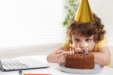 Photo of Birthday celebration. Little boy with cake having video call via laptop at table indoors. Space for text